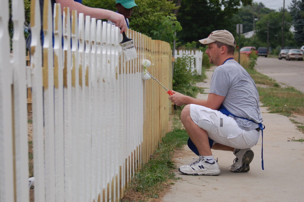 A man kneels on the sidewalk while painting a white picket fence