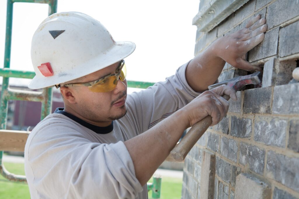 A man wearing a white hard head and safety goggles uses a hammer to repair a brick wall