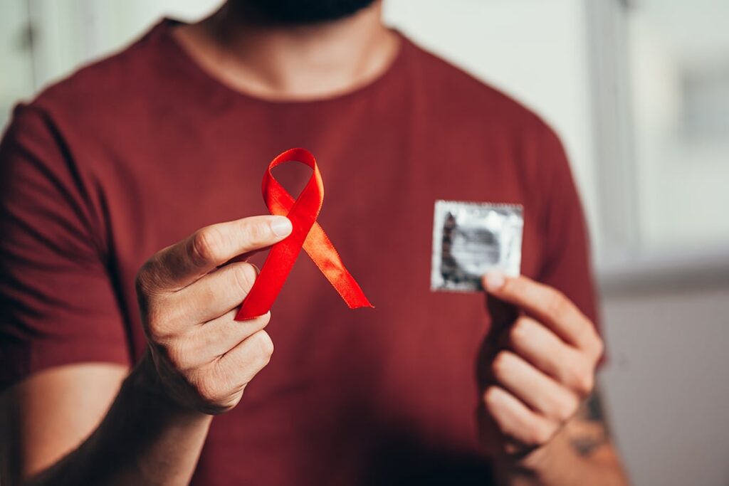 Person holding AIDS awareness ribbon in one hand and a condom in the other, promoting HIV/AIDS prevention.